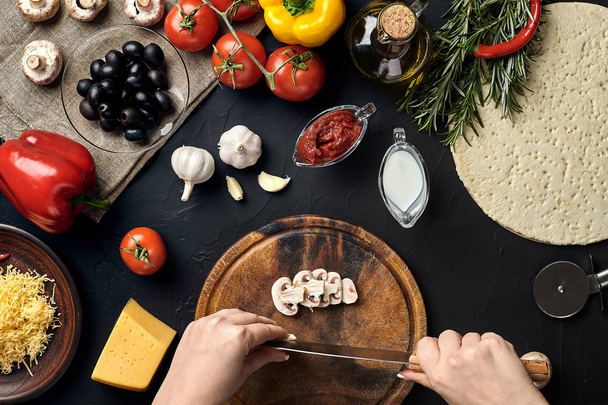 weibliche handgeschnittene Pilze auf Holzbrett auf Küchentisch, um sie herum liegen Zutaten für Pizza: Gemüse, Käse und Gewürze. - Foto, Bild