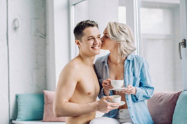girlfriend kissing boyfriend and holding cup of coffee at home - Foto, afbeelding