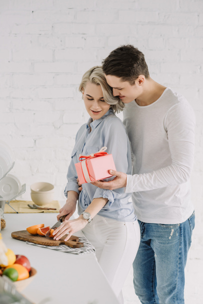 girlfriend cutting grapefruit and boyfriend gifting her present box in kitchen, international womens day concept - Foto, Imagen