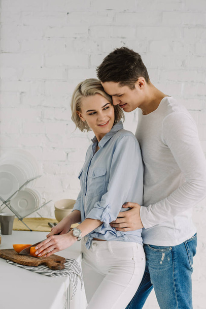 boyfriend hugging girlfriend cutting grapefruit in kitchen - Φωτογραφία, εικόνα