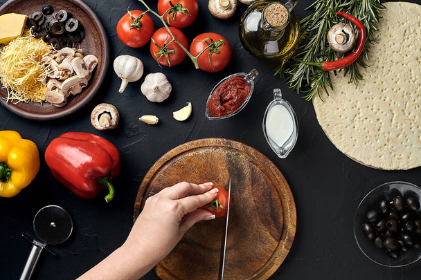 Female hand cut tomatoes on wooden board on kitchen table, around lie ingredients for pizza: vegetables, cheese and spices. - Photo, image