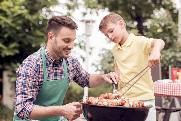 Freizeit, Essen, Menschen und Urlaub Konzept-Mann Kochen von Fleisch auf dem Grill mit seinem Sohn. - Foto, Bild