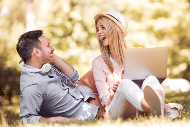 Młody piękny szczęśliwy uśmiechający się para siedzi razem w parku na letni dzień, za pomocą laptopa, śmiechu i zabawy. - Zdjęcie, obraz