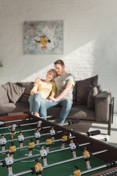 Paar umarmt und sitzt auf Sofa im Wohnzimmer zu Hause mit Tischkicker im Vordergrund - Foto, Bild