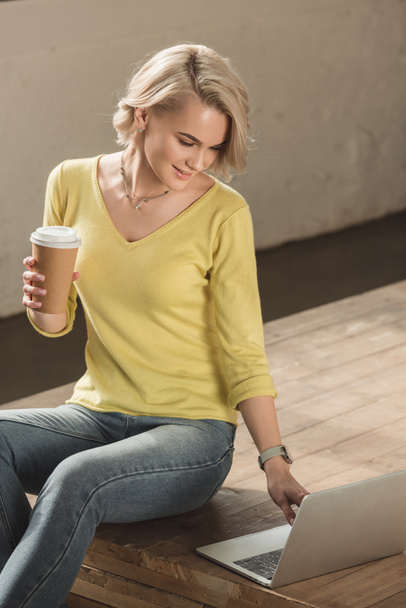 attractive girl holding coffee in paper cup and using laptop at home - Foto, immagini