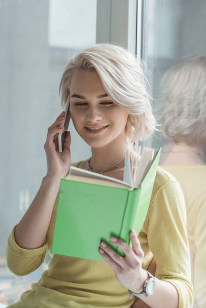 smiling girl talking by smartphone and reading book  - Φωτογραφία, εικόνα