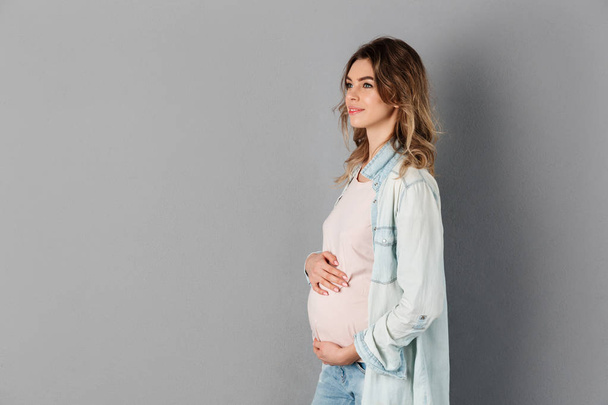 Visão lateral da mulher grávida sorridente segurando sua barriga
 - Foto, Imagem