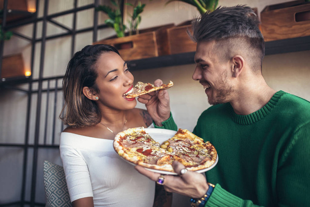 Pareja de razas mixtas comiendo pizza en un café moderno. Se ríen y comen pizza y se divierten
. - Foto, Imagen