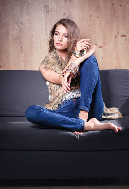 Portrait of elegant woman sitting on black sofa wearing a blue jeans and fur vest - 写真・画像