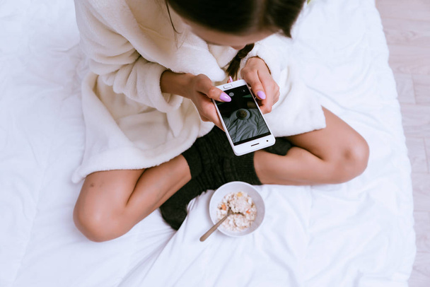 woman is sitting on the bed and holding a phone - Foto, afbeelding