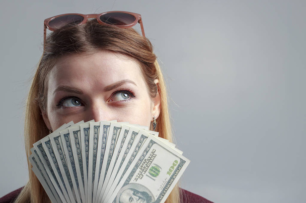 Een meisje in een Bourgondische sweatshirt heeft betrekking op haar gezicht met honderd-dollarbiljetten. - Foto, afbeelding