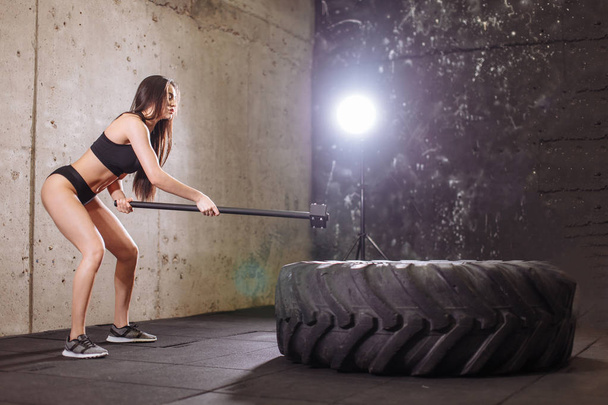woman smashing large tire with sledgehammer during intense workout in fit gym - Photo, image