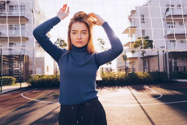 stylish beautiful blond girl in a blue sweater posing on a football field outdoors in the sun - Фото, изображение