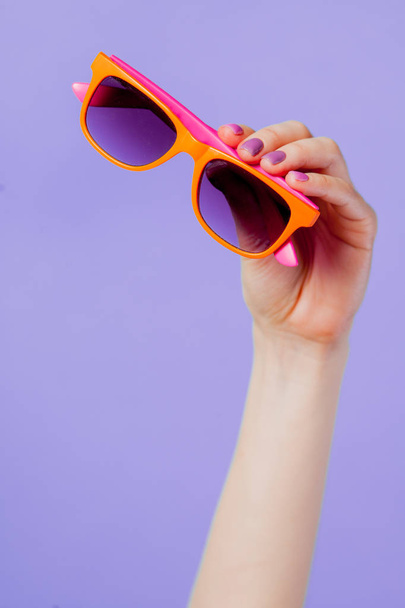 Female hand holding orange eyeglasses on purple background - Foto, immagini