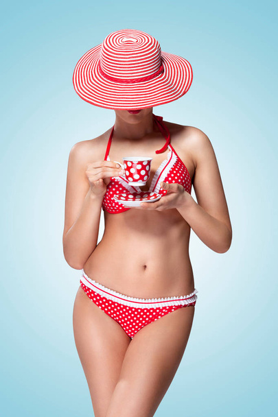 A vintage photo of a beautiful pin-up girl in bikini drinking tea and hiding head under a sun hat. - 写真・画像