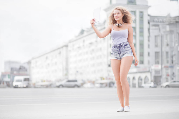Happy young woman with vintage music headphones around her neck, taking selfie with tablet pc and smiling happily against urban city background. - Fotó, kép