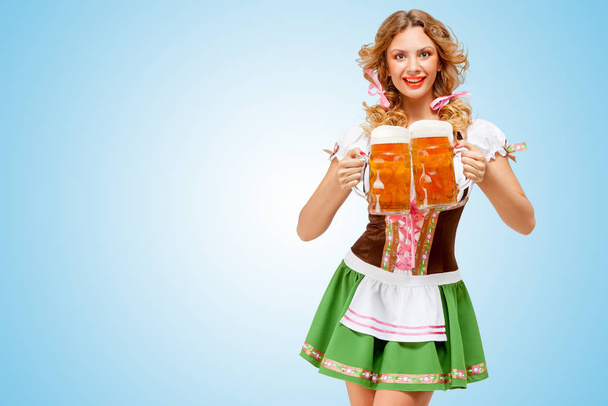 Young sexy Oktoberfest woman wearing a traditional Bavarian dress dirndl serving two beer mugs with happy smile on blue background. - Foto, Imagem