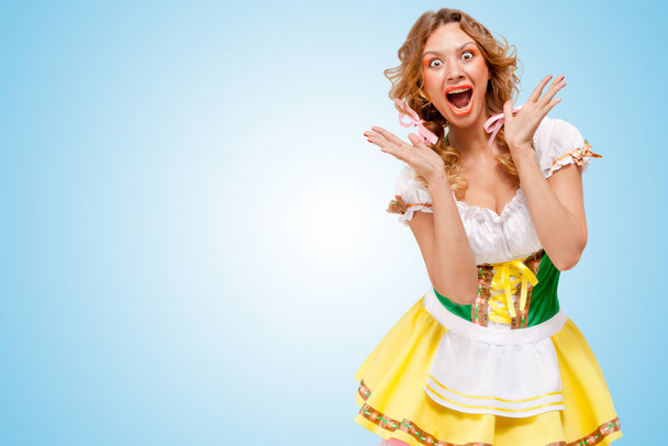 Young sexy astonished Oktoberfest woman wearing a traditional Bavarian dress dirndl, showing happy emotions on blue background. - 写真・画像