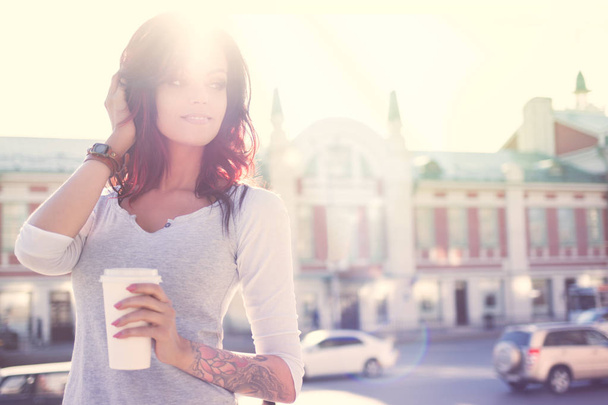 mooie jonge lachende vrouw met tatoeage, houden een wegwerp afhaalmaaltijden kopje en opzij op zoek tegen stedelijke stad achtergrond. - Foto, afbeelding