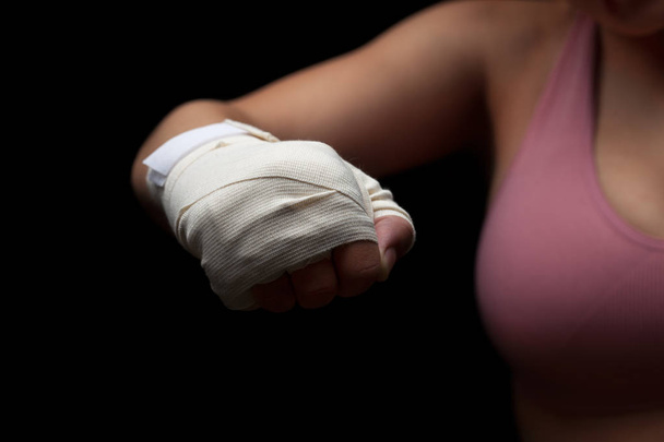 Beautiful and fit female fighter getting prepared for the fight or training, holding wrapped fist ready for punch against dark background. - Fotó, kép