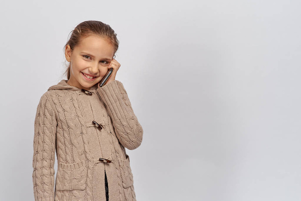 little young lady in a warm brown sweater looking at camera and holding telephone, child communicates in a pleasant - Photo, image