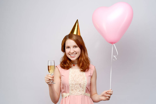 lovely European girl with red hair and a festive hat widely welcoming smiles. She holds in her hands a glass and a balloon  - Фото, изображение