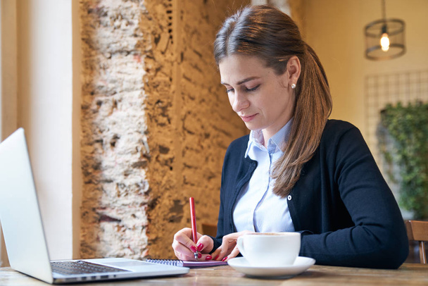felnőtt női dolgozó egy asztalnál, jegyzetelés egy füzetbe egy csésze kávé és laptop kávézóban üzleti portré - Fotó, kép