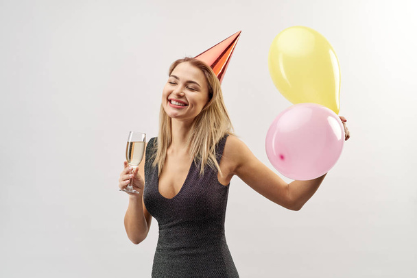 portrait of attractive young woman in a dark dress with a glass of champagne, festive cap on her head and balloons - Foto, Bild