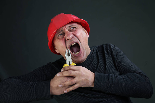 portrait of a man in a black shirt and red hard hat, rips a tooth with pliers studio - Fotografie, Obrázek