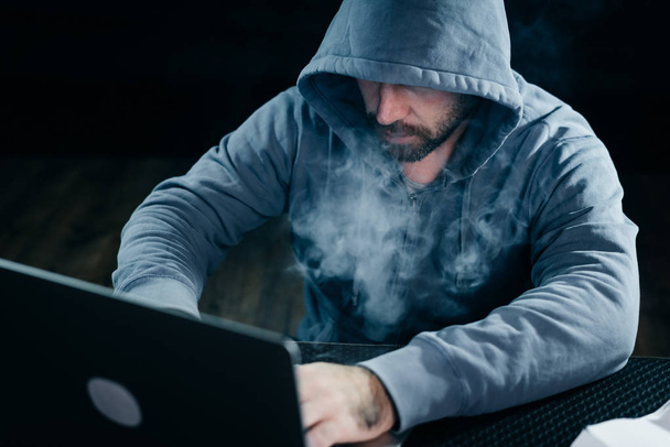 a criminal man hides his face under a hood, hacks a laptop, lots of smoke - 写真・画像