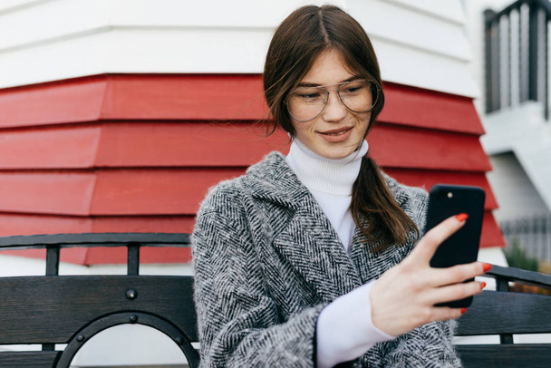 a nice dark-haired girl with glasses and a gray coat is sitting on a bench in the open air, making selfies - Foto, Imagen