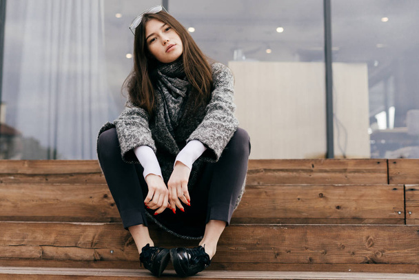 stylish dark-haired city girl in gray coat sits on steps, posing and looking at camera - Valokuva, kuva