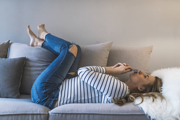 Smiling young woman at home relaxing on the couch, she is using a smartphone and texting, technology and communication concept. Pretty girl using her smartphone on couch at home in the living room. Photo of a beautiful young woman in sofa with smartp - 写真・画像