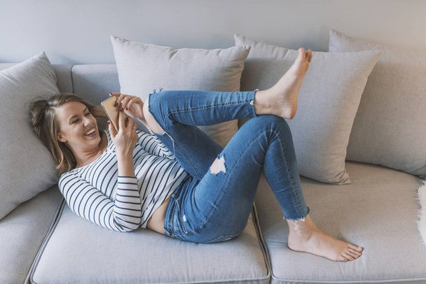 Smiling young woman at home relaxing on the couch, she is using a smartphone and texting, technology and communication concept. Pretty girl using her smartphone on couch at home in the living room. Photo of a beautiful young woman in sofa with smartp - Фото, изображение