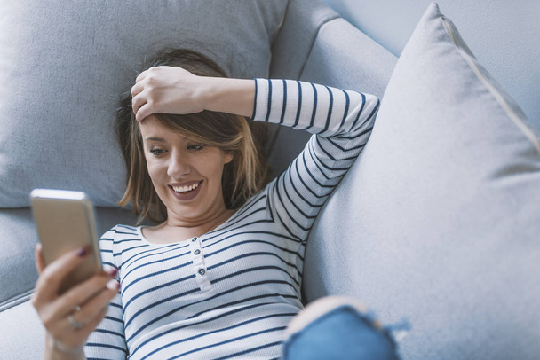 Girl reading message in a smart phone. Happy woman using mobile phone on sofa. Happy woman at home reading a text message in her bright livingroom. Relaxed young woman text messaging on sofa - Photo, Image