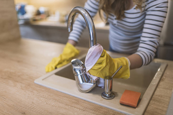 people, housework and housekeeping concept - happy woman in protective gloves cleaning tap with rag at home kitchen. Close-up of woman wiping kitchen sink with rag as cleanliness concept. Woman washing the kitchen sink and faucet. Happy Woman Cleanin - Foto, afbeelding