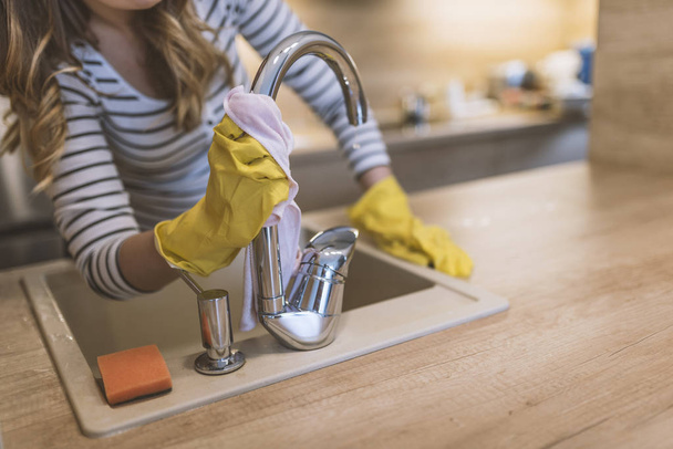 people, housework and housekeeping concept - happy woman in protective gloves cleaning tap with rag at home kitchen. Close-up of woman wiping kitchen sink with rag as cleanliness concept. Woman washing the kitchen sink and faucet. Happy Woman Cleanin - Zdjęcie, obraz