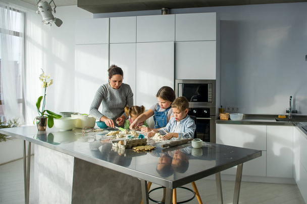 Mother teaches her three children to cook. Family is preparing something from the dough. There are necessary products on the kitchen table. Useful and fun pastime. - Foto, Imagen