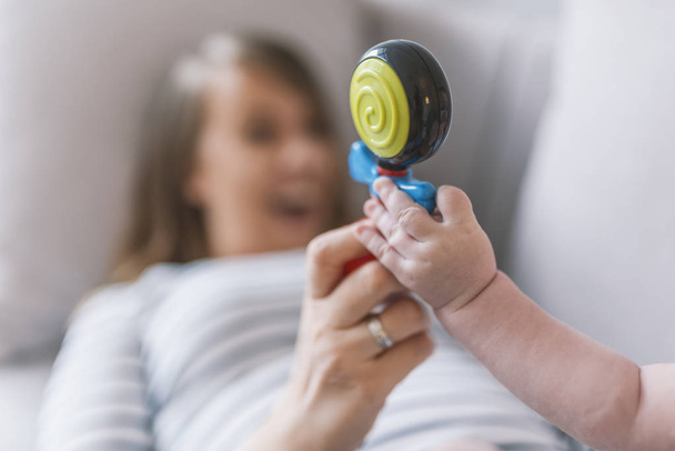 Mother play with baby with rattle. Family concept photo, lifestyle, view above, focus on a toy. Beautiful mom holding her baby in arms with rattle. Portrait of a happy female and her toddler son playing with rattle - Foto, immagini