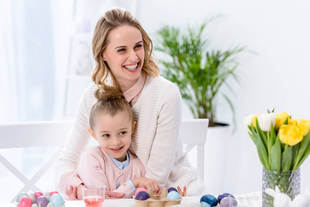 Весела мати і дочка за столом з розфарбованими яйцями на Великдень
 - Фото, зображення