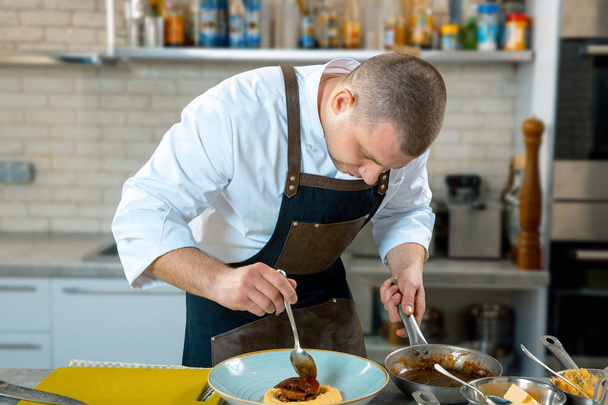 food cooking, profession and people concept - male chef cook serving plate of polenta and veal tongues with sauce at restaurant kitchen. cooking process - Photo, Image