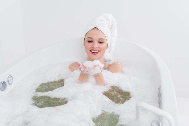 femme souriante avec serviette sur la tête relaxant dans la baignoire avec mousse dans le salon de spa
 - Photo, image