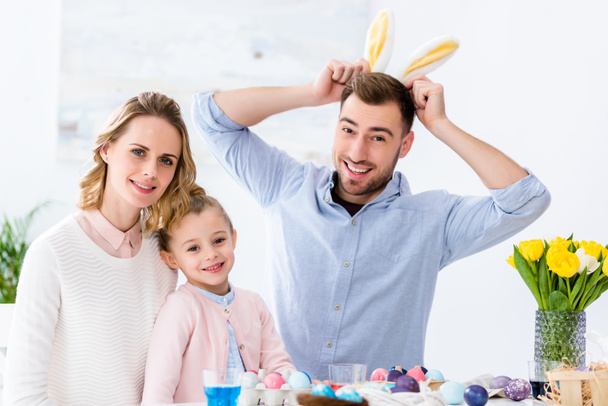 Happy family playing with bunny ears by table with colored eggs for Easter - Фото, зображення