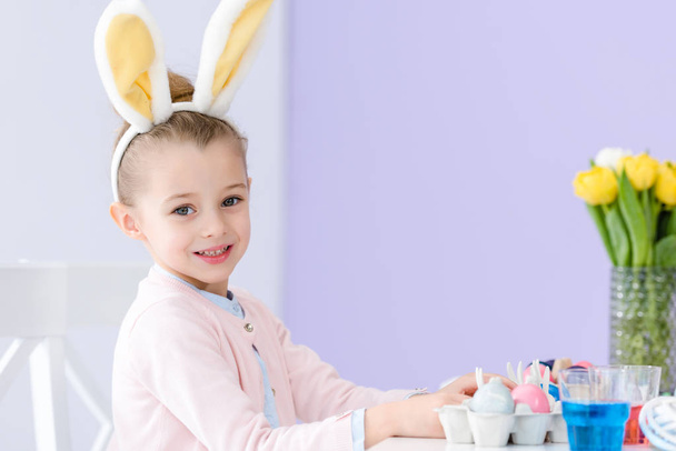 Child girl playing wearing bunny ears by table with Easter colored eggs - Foto, afbeelding