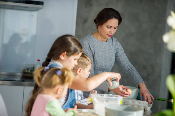 Mother teaches her three children to cook. Family is preparing something from the dough. There are necessary products on the kitchen table. Useful and fun pastime. - Fotografie, Obrázek
