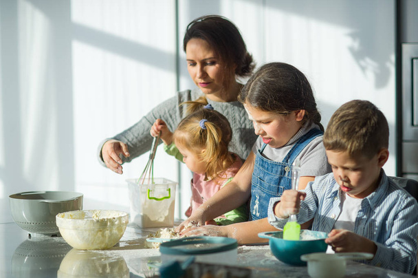 Мати вчить своїх трьох дітей готувати. Сім'я щось готує з тіста. На кухонному столі є необхідні продукти. Корисна і весела розвага. - Фото, зображення
