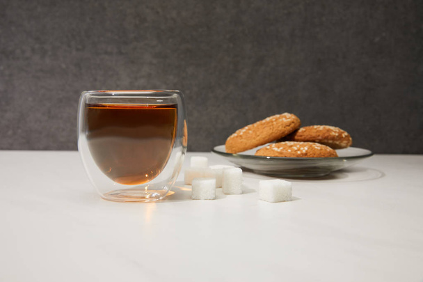 Nahaufnahme von frischem heißen Tee in Glasschale, Zuckerwürfeln und leckeren Keksen auf grau  - Foto, Bild