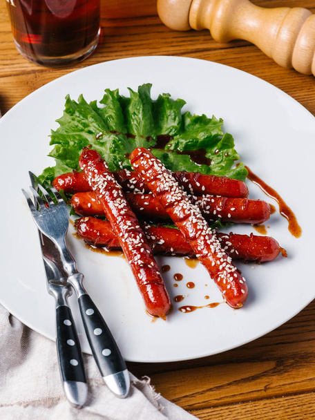 Жареные охотничьи сосиски с кунжутом и салатом подаются на белой тарелке с ножом, вилкой, темным пивным бокалом и мясорубом на деревянном столе
 - Фото, изображение