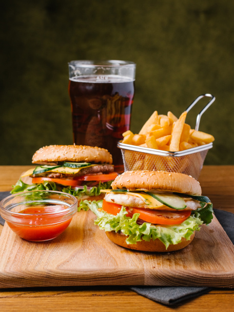 Food composition of cheeseburgers served with french fries in basket, dark beer and sweet ketchup on wooden chopping board and grey napkin - Foto, Bild