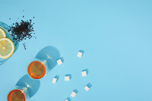 вид зверху на свіжий чай в чашках, цукрові кубики, сухий трав'яний чай і нарізаний лимон на синьому
  - Фото, зображення